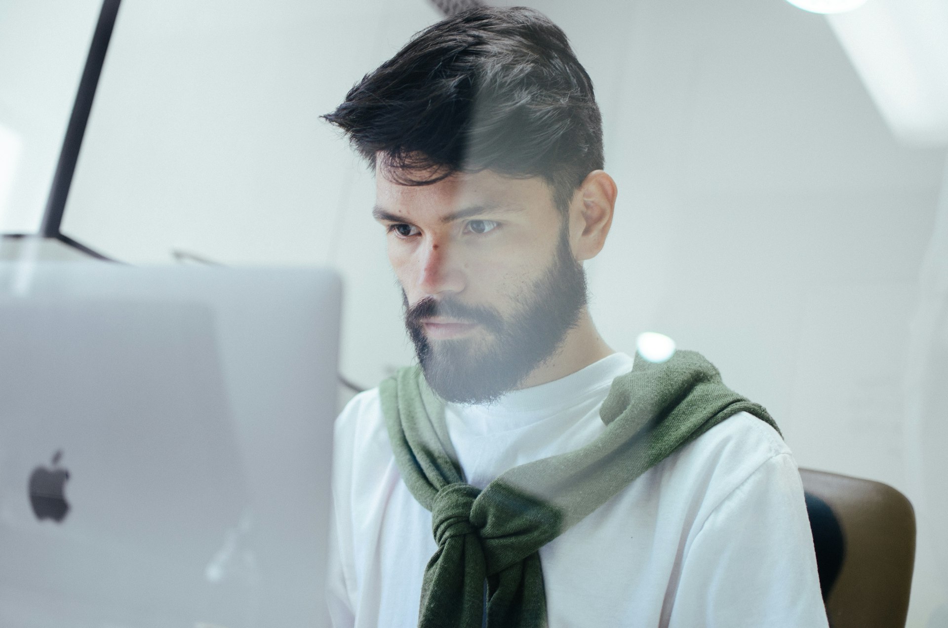 Professionnel Concentré Travaillant sur un Ordinateur Portable dans un Bureau Moderne Jeune professionnel concentré avec une barbe travaillant sur un ordinateur portable dans un bureau moderne, vêtu d'un t-shirt blanc et d'un pull vert sur les épaules.